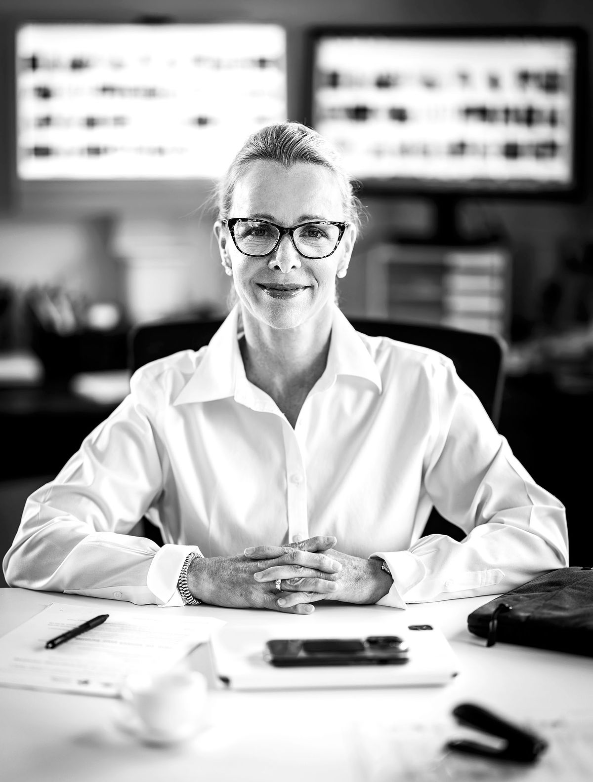 Corporate portrait of Louise. A CEO of a medium size company. Sitting at her busy desk. Computers in the background.