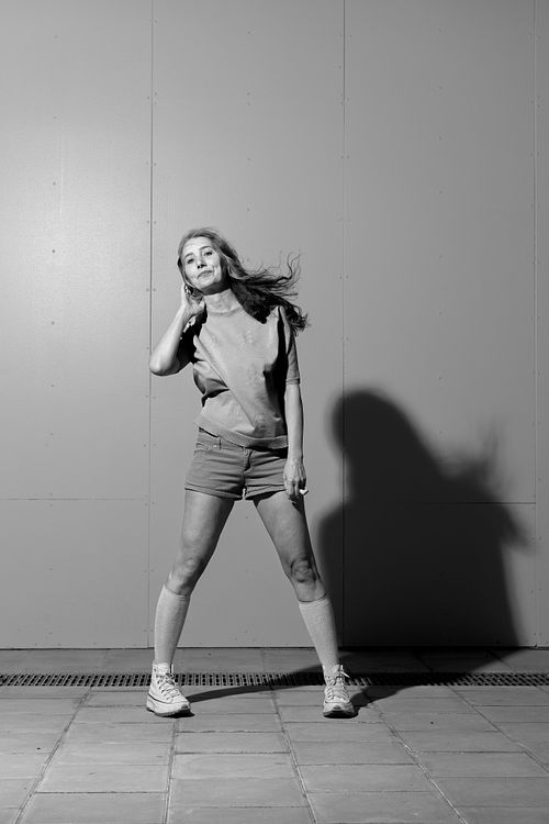 Black and white portrait of a woman in shorts standing against a plain wall with dramatic shadow, photographed in Stockholm by Mats Karlsson.