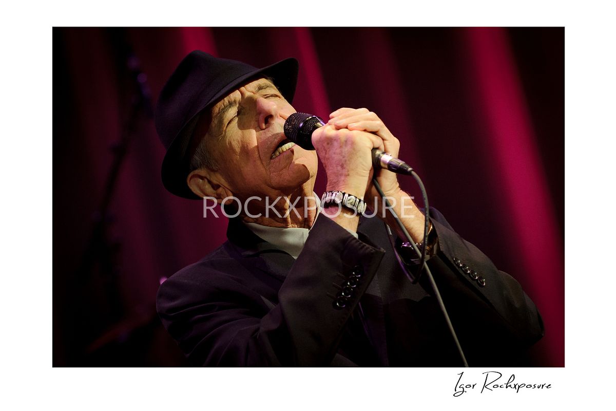 Horizontal concert photography of Leonard Cohen singing into a microphone under red stage lighting