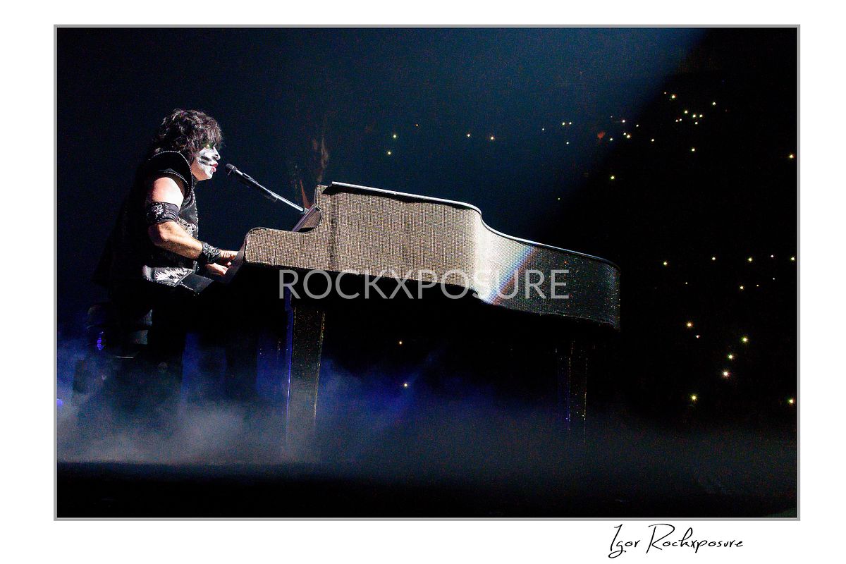 Horizontal color photograph of Eric Singer in full KISS makeup and costume seated at a glittering grand piano while singing under soft concert lighting with scattered lights visible in the background