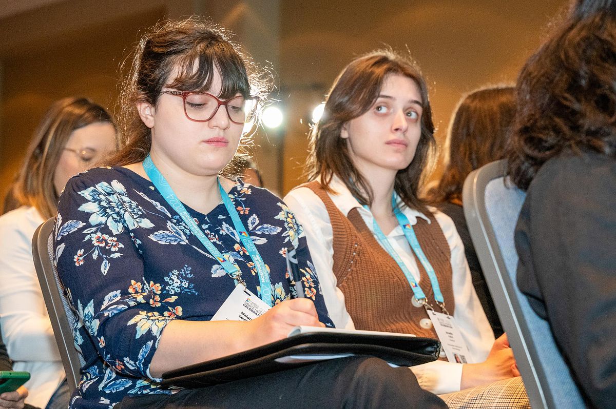 Two conference attendees seated and attentive, one writing notes during a corporate event keynote session.
