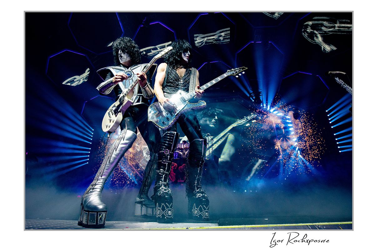 Horizontal color photograph of Paul Stanley and Tommy Thayer performing side by side in full makeup and costumes with bright stage lighting, hexagonal screens, and pyrotechnics behind them