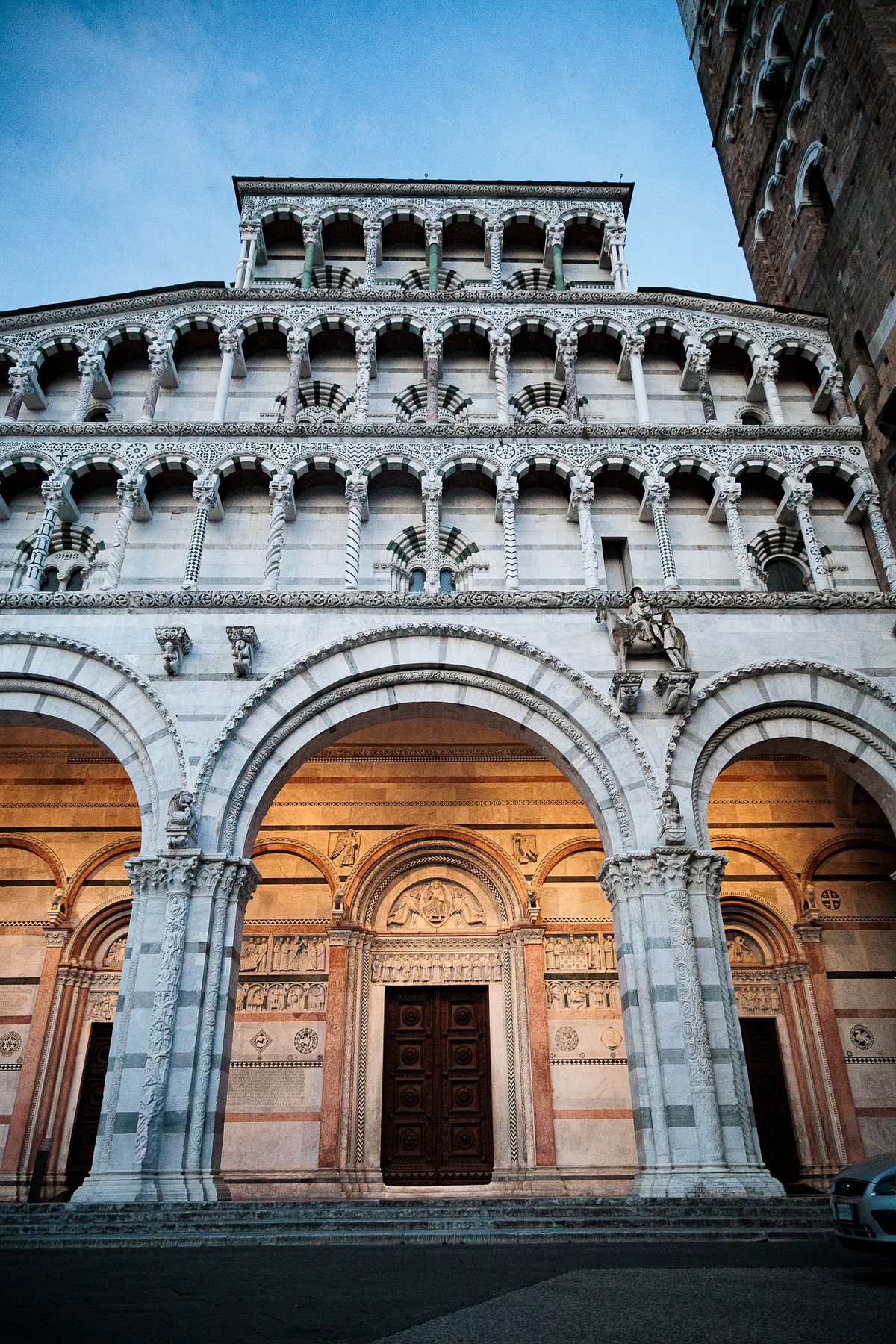 Evening view of Lucca Cathedral&rsquo;s ornate stone facade in Italy, showing multiple layers of arches and columns, a large wooden entrance door framed by carved reliefs, and contrasting light and dark stone patterns, softly lit against a clear blue sky with part of the street and a vehicle visible in the foreground.