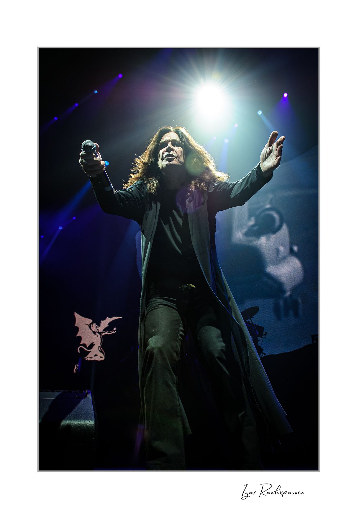 Ozzy Osbourne of Black Sabbath standing with both arms raised on stage under dramatic blue concert lighting during a live performance