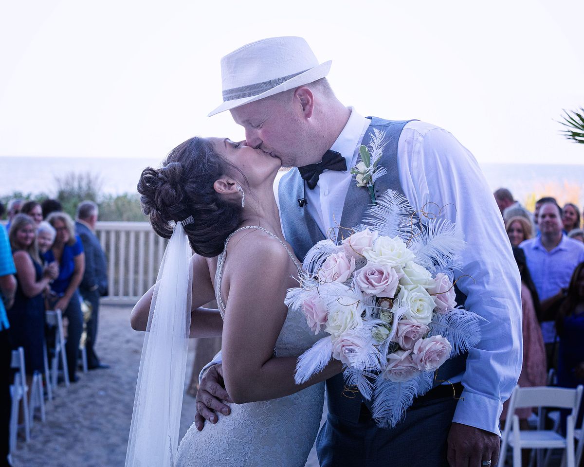 a bride and groom kissing after their wedding ceremony at Golden Sands in Ocean City. Her flower bouquet is modelled after the Great Gatsby, with pastel coloured roses and feather