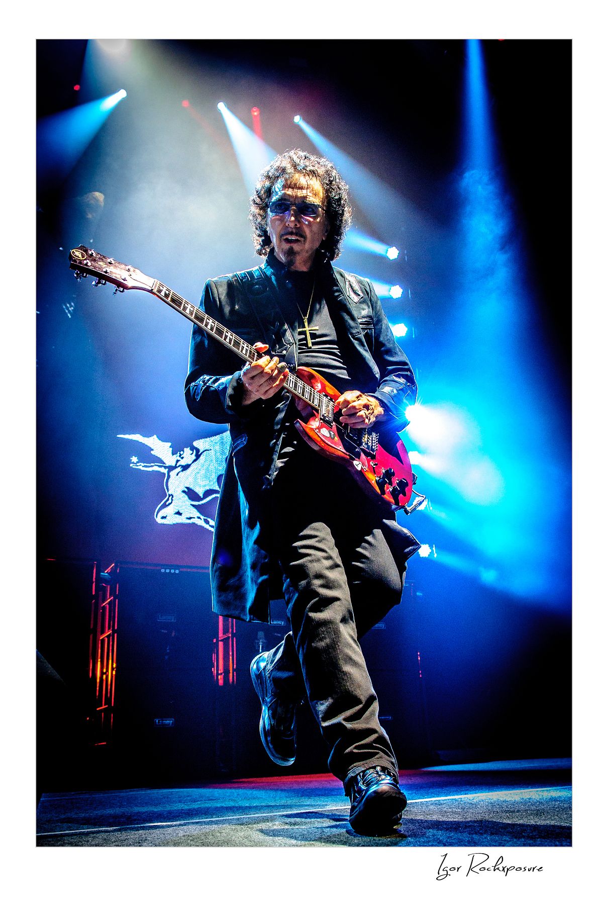 Tony Iommi of Black Sabbath walking across the stage while playing electric guitar under dramatic blue concert lighting
