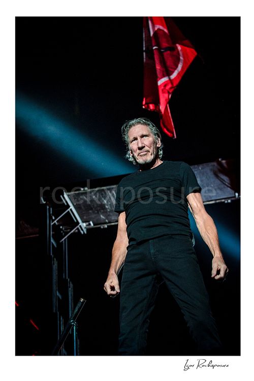 Vertical color image of Roger Waters on stage in a dramatic live performance pose, standing beneath a red flag with a strong spotlight cutting across the dark background