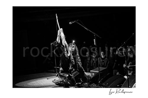 Horizontal black and white image of Yngwie Malmsteen kneeling on stage and lifting his white guitar high beside the microphone, with the crowd watching from just beyond the monitors