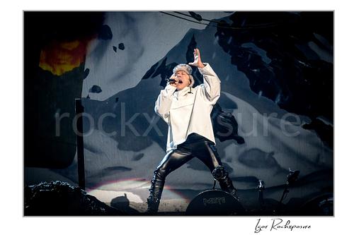 Bruce Dickinson of Iron Maiden performing live on stage in a white outfit, singing into a microphone with one hand raised against a dramatic dark stage backdrop.