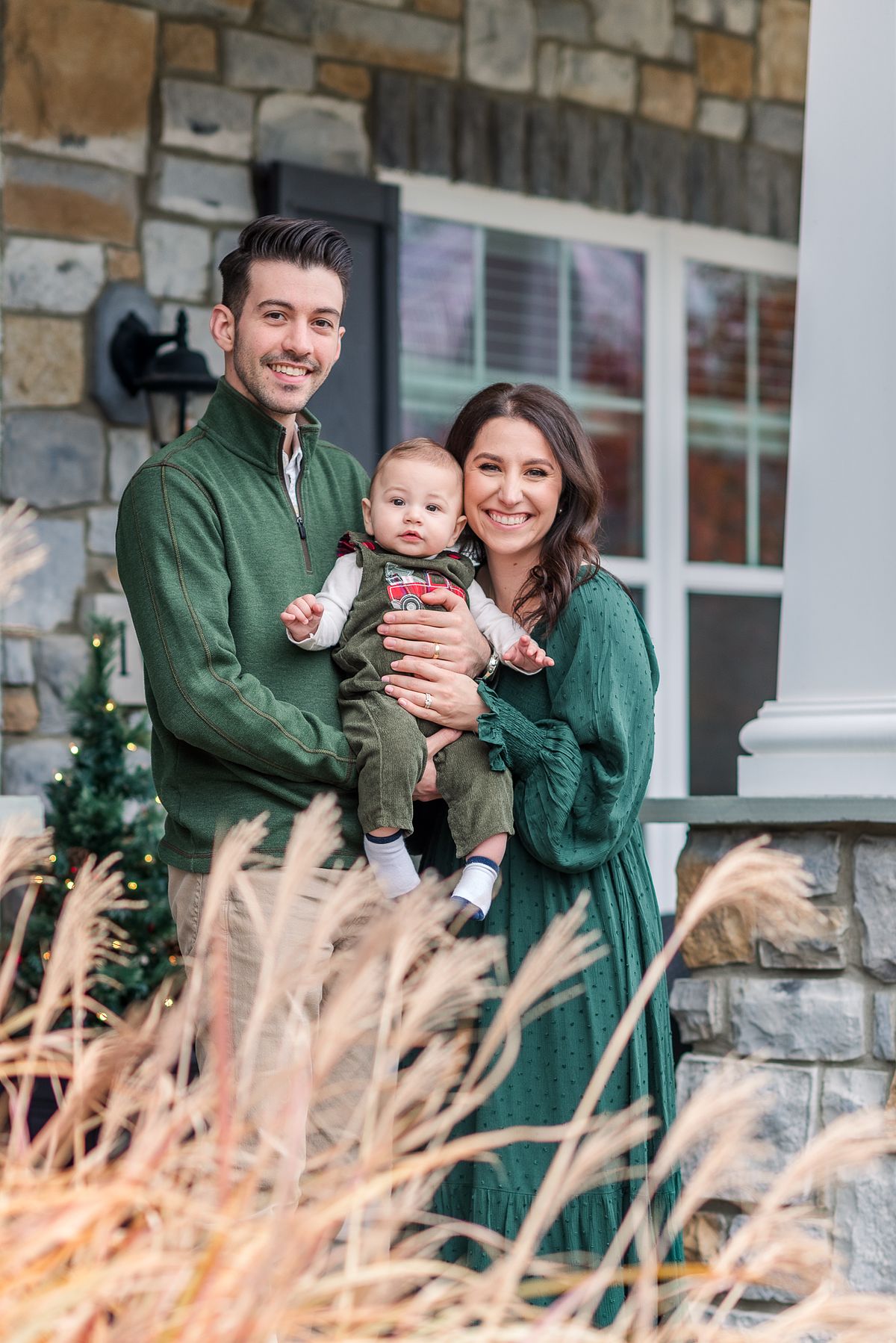 Husband and wife smiling and holding baby on porch dressed for Christmas with Cranberry Township, PA newborn photographer