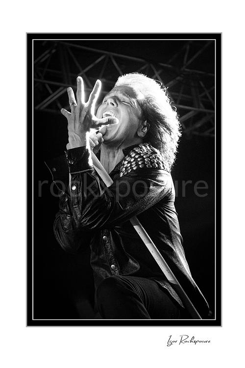 Vertical black and white image of Joey Tempest of Europe performing live on stage with a white microphone stand, studded black jacket, bright backlight, and expressive singing pose