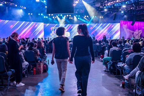 Two attendees walk hand-in-hand toward the main stage at the Grace Hopper Celebration 2024, with vibrant lighting and a full crowd at this landmark tech event for women and non-binary technologists.
