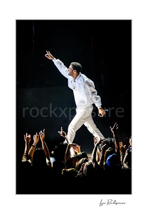 Vertical color image of Drake performing live on stage in a white hoodie and white pants with a handheld microphone, raised arm, and fans reaching up from the crowd