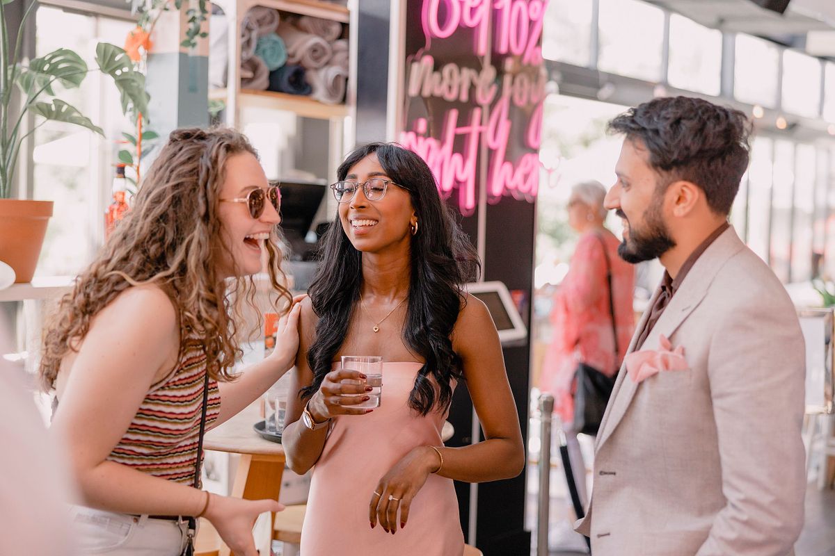 Photographer in Basel capturing three friends chatting at an engagement party and laughing in a lively caf&eacute; setting. One holds a drink, with a neon sign in the background, creating a joyful atmosphere.