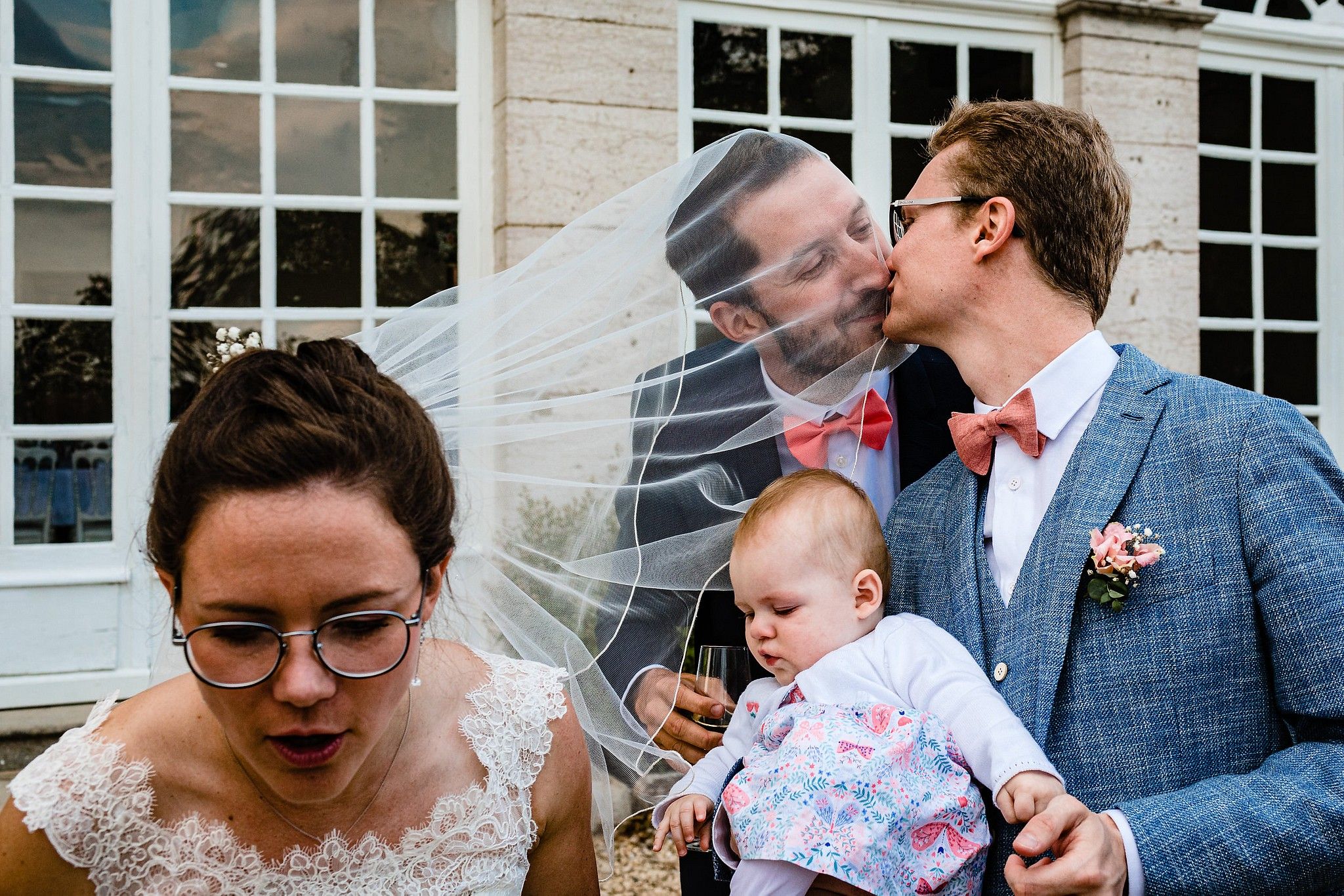 Couple de Mari&eacute;s qui s'amusent. Le t&eacute;moin du mari&eacute; vient l'embrasser en mettant sa t&ecirc;te sous le voile de la mari&eacute;e captur&eacute; par S&eacute;bastien CLAVEL photographe de Mariage &agrave; Lyon et Gen&egrave;ve