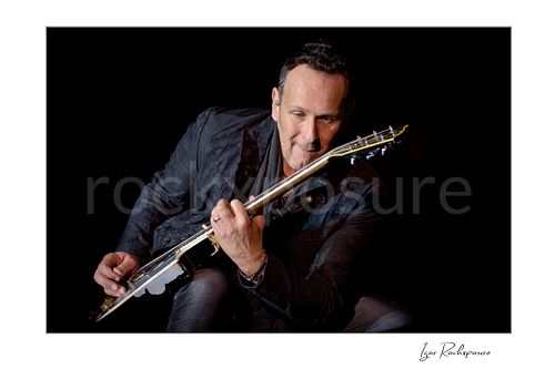 Horizontal color image of Vivian Campbell of Def Leppard performing live on stage, playing electric guitar against a dark background