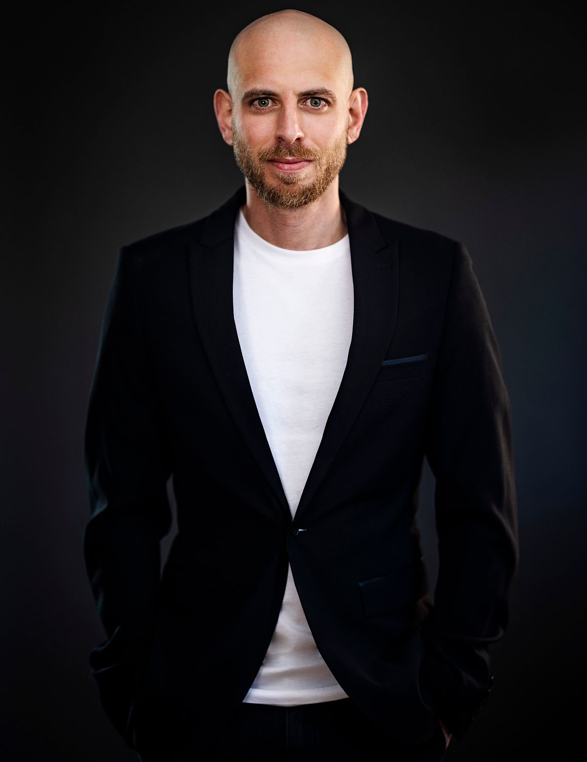 Corporate portrait against a dark background. A man wearing a dark suit and white tshirt.