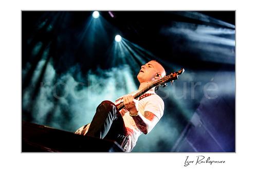 Horizontal color image of Johnny Christ of Avenged Sevenfold playing bass onstage beneath bright concert beams and drifting smoke.
