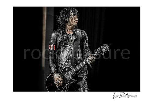 Horizontal color image of Tom Keifer performing live on stage with a black guitar, wet hair falling across his face against a dark concert background