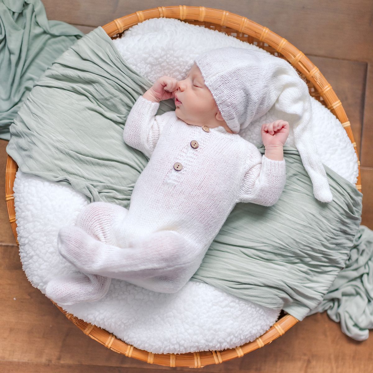 Baby boy in white pajamas and hat sleeping in a circular basket with fluffy blankets and green accent