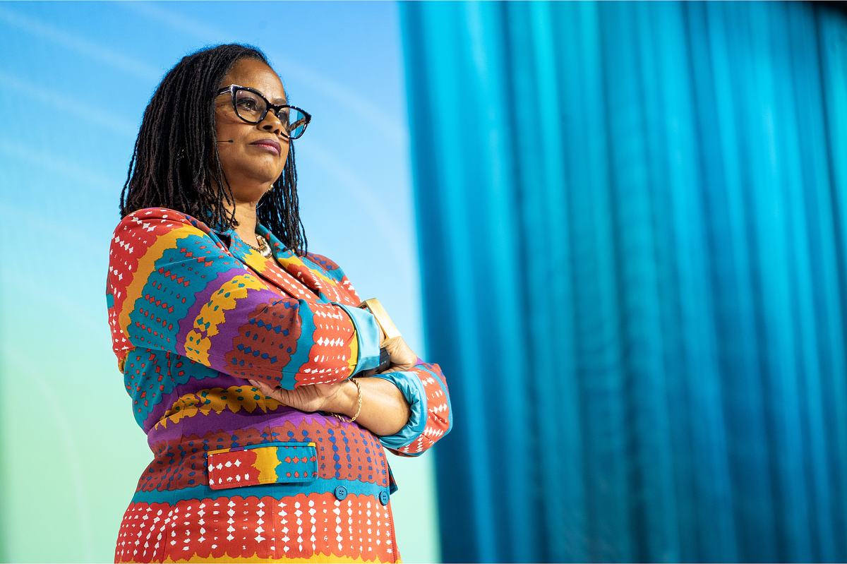 Corporate event photography capturing a keynote speaker standing confidently on stage during a major leadership and technology conference, wearing a bold patterned suit against a vibrant blue backdrop, emphasizing representation, presence, and impact.