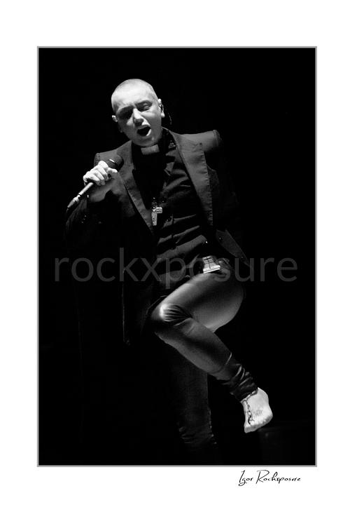 Vertical black and white image of Sinéad O’Connor singing into a microphone on stage in a dramatic low-light portrait