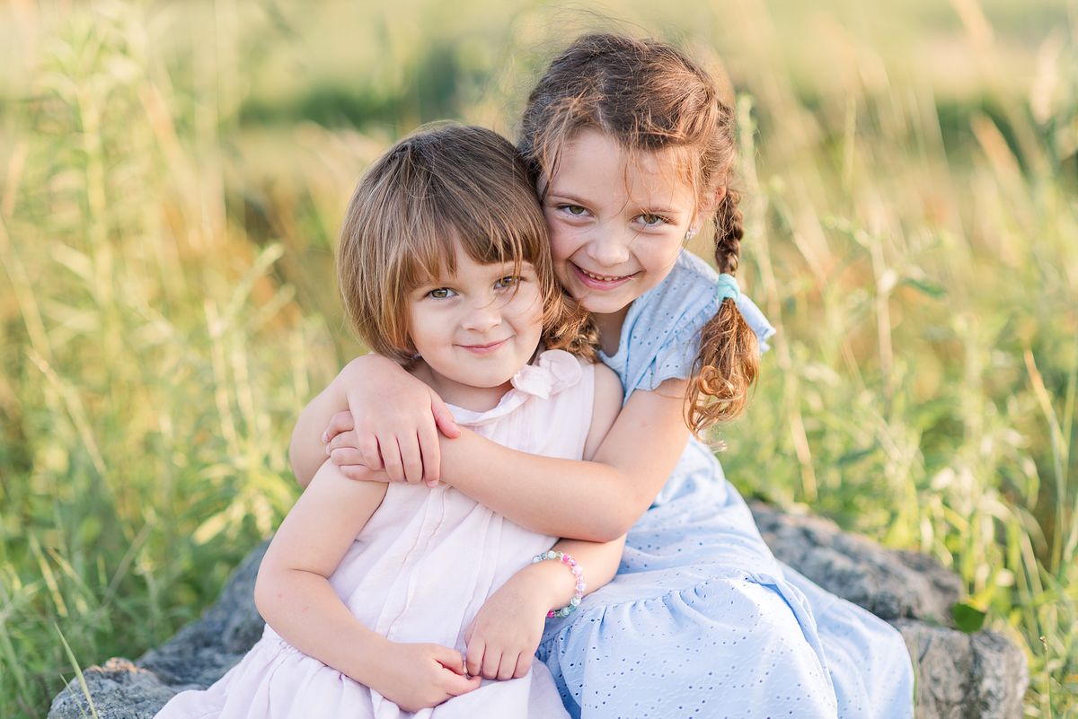 Two sisters hugging on a large rock in a sunset lit grassy field at Cranberry Township Community Park