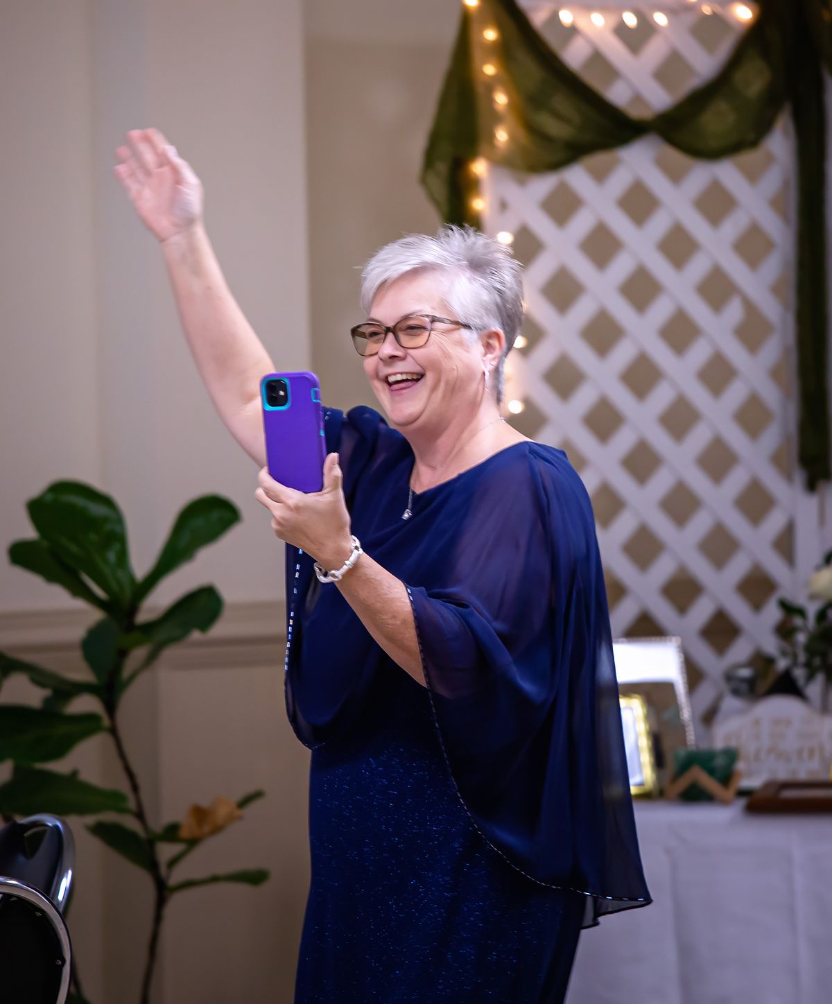 A candid, documentary-style shot of a wedding guest in a navy blue dress laughing and waving while holding a phone at a reception.