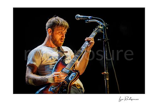 Horizontal color image of Dan Auerbach of The Black Keys playing guitar beside a microphone under warm stage lighting