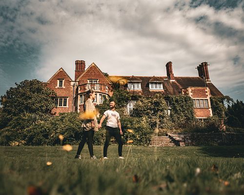 Alex and Shelby holding hands in Oadby Botanical Gardens, with a beautiful historic building in the background, captured by Weddings by Jermaine during their engagement shoot