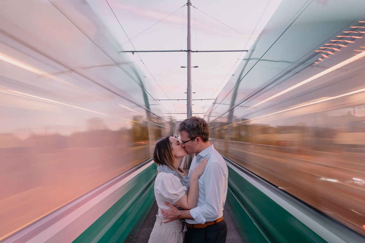 A Basel couple kisses tenderly between iconic green Basel trams, with blurred motion of two passing trains framing them, under a soft pink sunset sky. Romantic and dynamic.