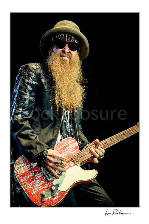 Vertical color image of Billy Gibbons of ZZ Top performing live on stage with a red Telecaster guitar, wearing a wide-brim hat, dark glasses, and his signature long beard against a dark background