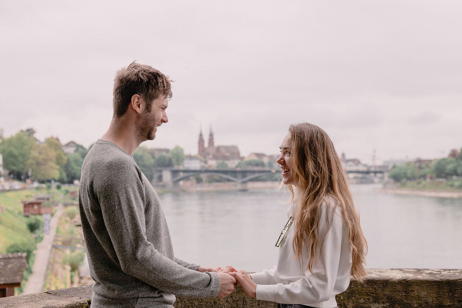 Basel photographer capturing a couple holds hands, smiling at each other by the Rhine river with a bridge and historic Munster cathedral in the background. The scene is serene and romantic.